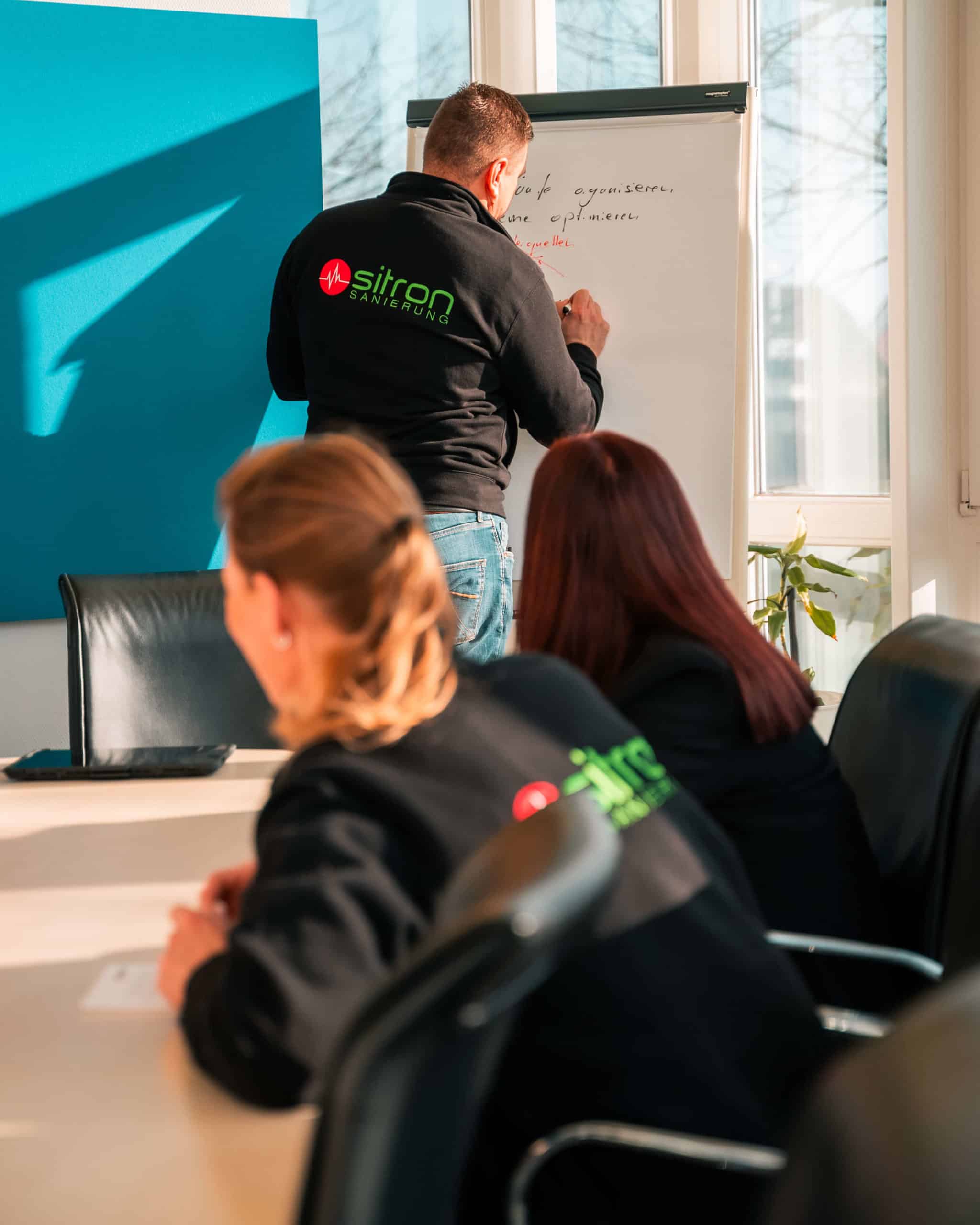 Vorarbeiter von Sitron steht am Flippchart und erklärt im Besprechungsraum zwei Mitarbeiterinnen Details über einen Auftrag.