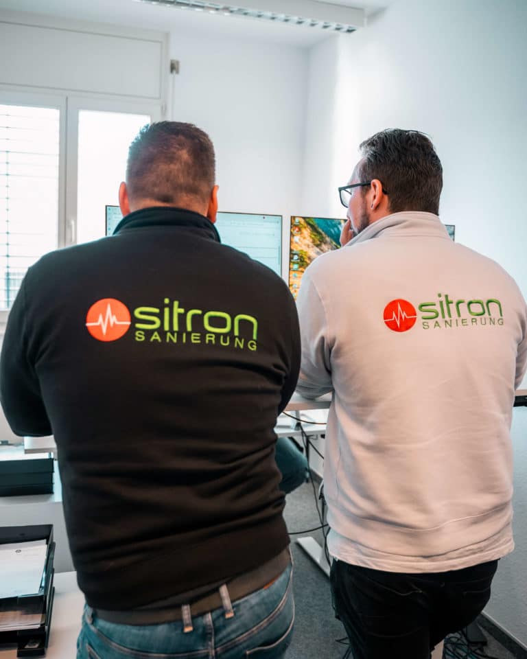 Zwei Mitarbeiter von Sitron mit Arbeitskleidung stehen an einem Schreibtisch und schauen gemeinsam auf Computerbildschirme, um einen Auftrag zu besprechen.