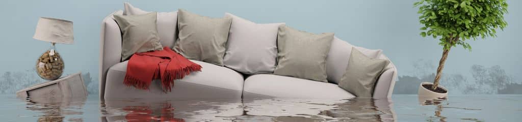 Großes, helles Sofa mit Kissen schwimmt im Wasser. Links daneber schwimmt ein Beistelltisch mit einer Lampe, rechts davon schwimmt eine Pflanze mit Blumentopf.